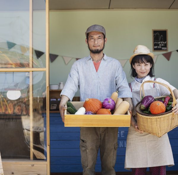 Japanese man and woman standing outside a farm shop, holding crate and basket with fresh vegetables, looking at camera.