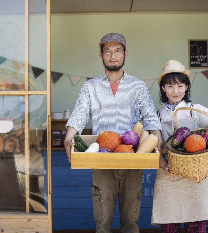 Japanese man and woman standing outside a farm shop, holding crate and basket with fresh vegetables, looking at camera.