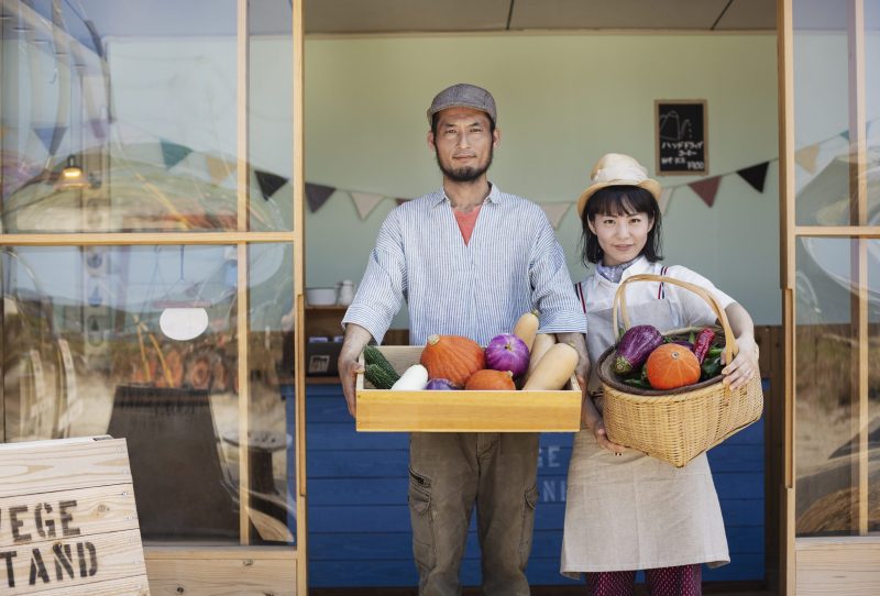 Japanese man and woman standing outside a farm shop, holding crate and basket with fresh vegetables, looking at camera.