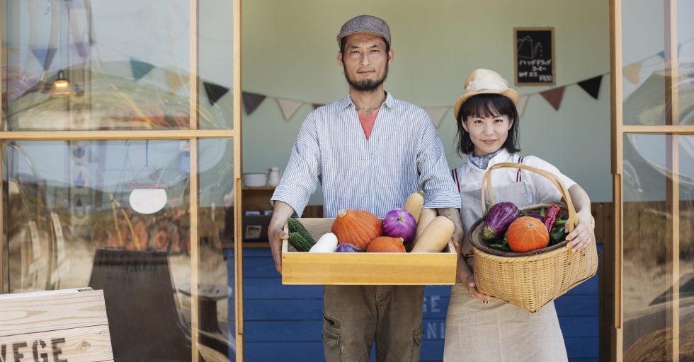 Japanese man and woman standing outside a farm shop, holding crate and basket with fresh vegetables, looking at camera.